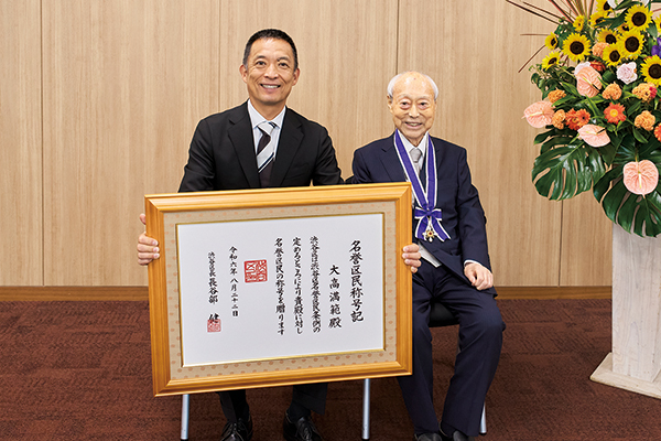 名誉区民称号記を前に並ぶ大高さんと長谷部区長の写真。大高さんは首から名誉区民証をさげている
