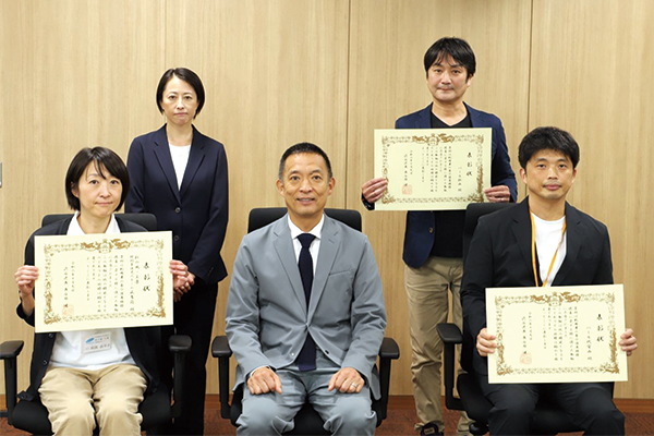 長谷部区長と表彰状を掲げる事業所の皆さんの記念写真